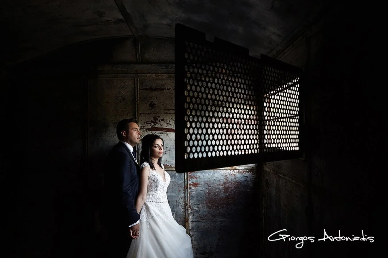 A bride and groom standing together indoors, with the groom in a dark suit and tie, and the bride in a white wedding dress, holding hands, looking to the side, in front of a dark, industrial-style wall and a large window with circular holes, allowing