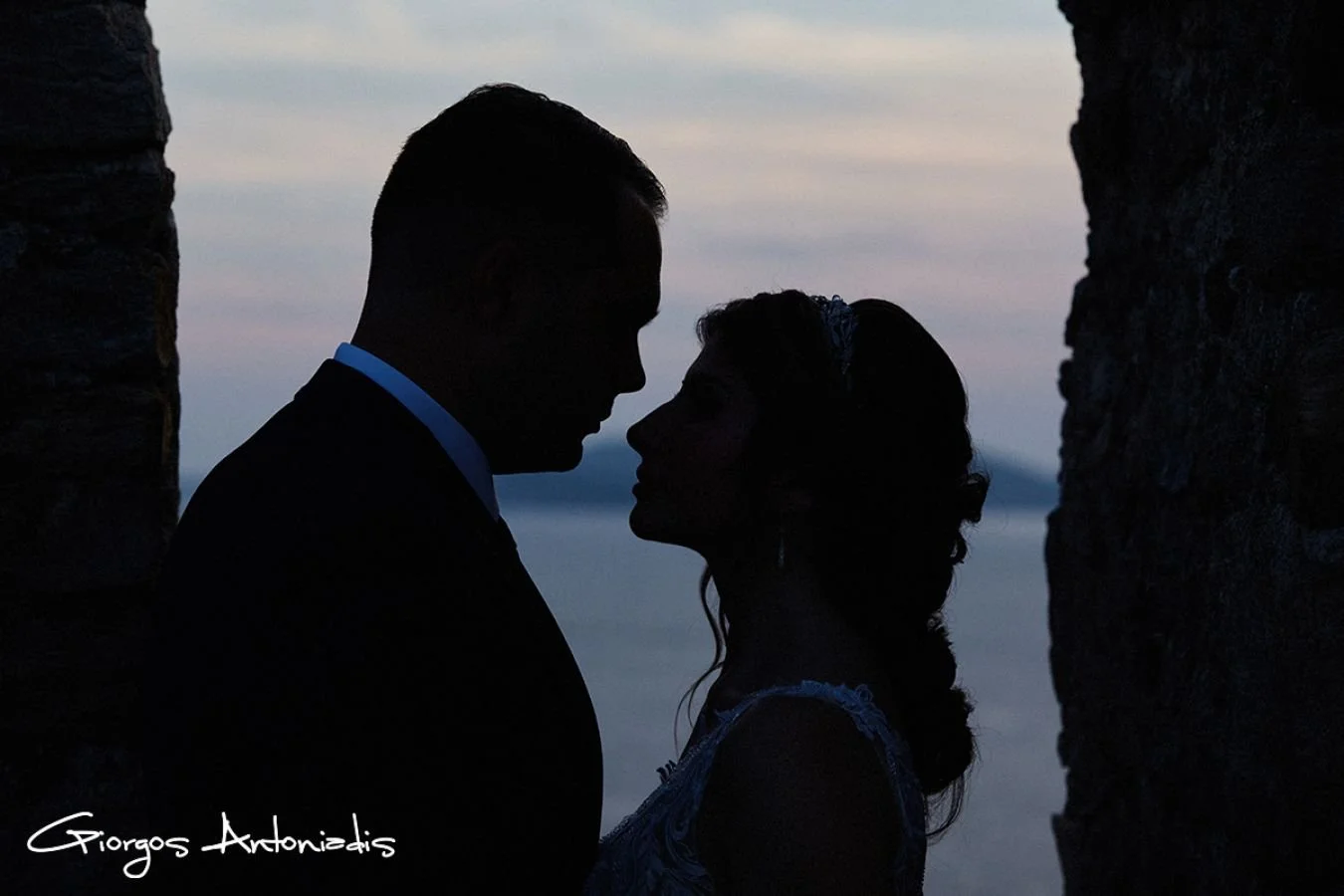 Silhouette of a couple facing each other, close together, between two stone walls, during sunset or dusk.