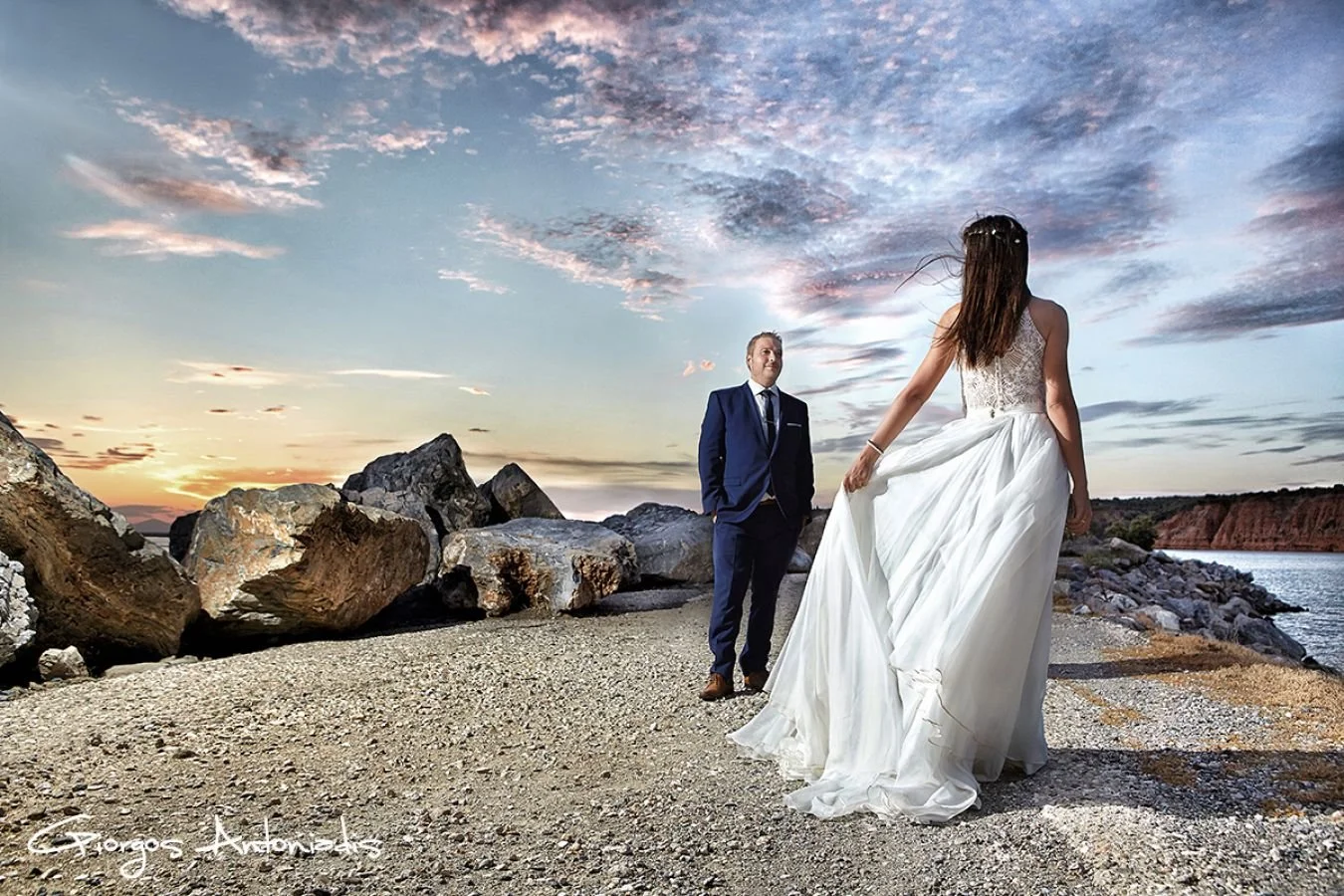 A bride in a white wedding dress is walking towards a groom in a navy suit on a rocky beach at sunset, with a colorful sky and water in the background.