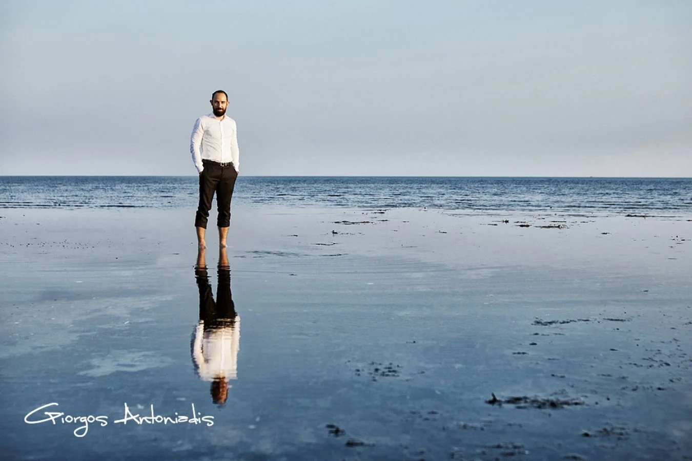 A man standing in shallow waters at the beach with a calm ocean and clear sky in the background, his reflection visible in the water. The photo is signed by Giorgos Antoniadis.