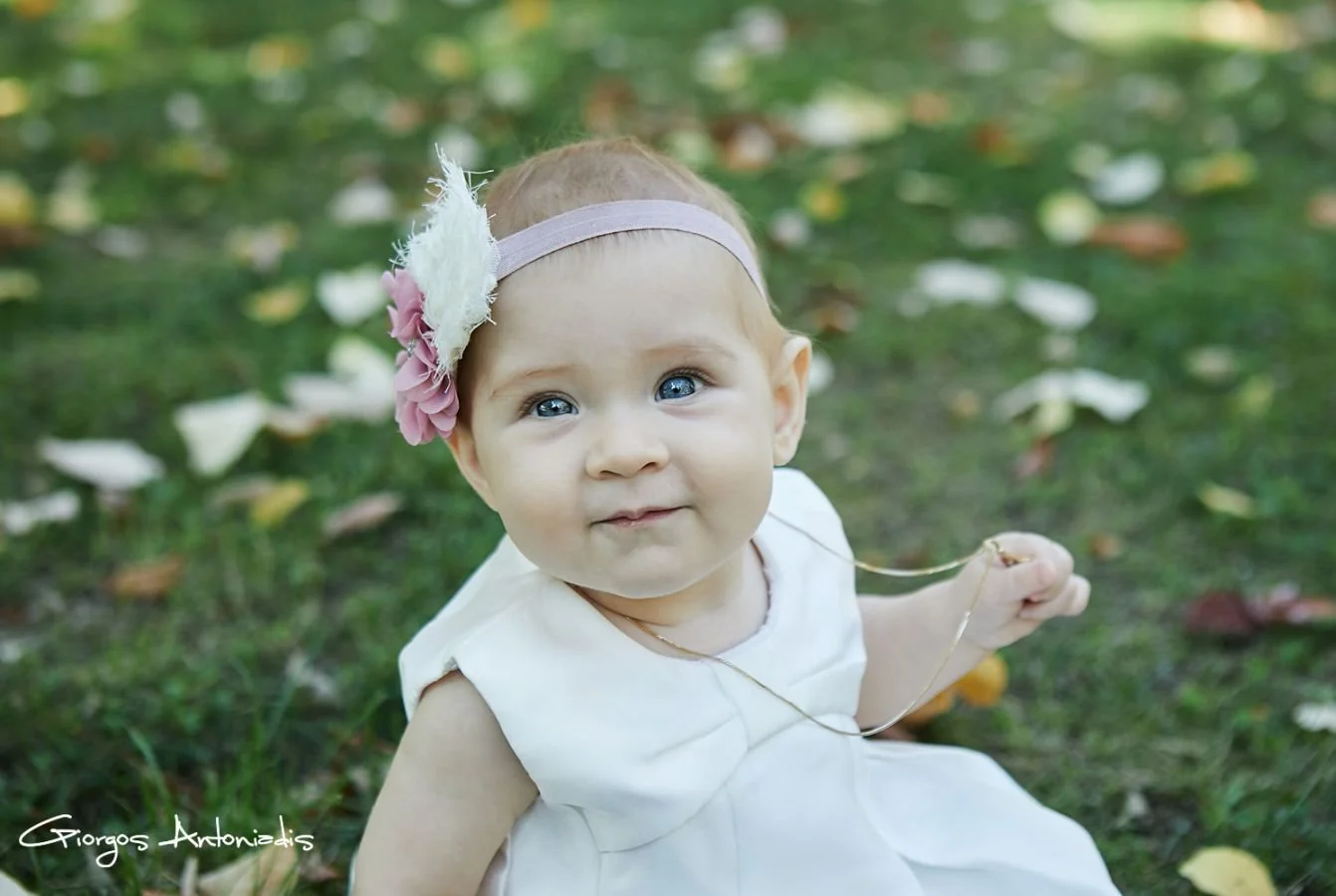 A close-up of a baby girl with blue eyes sitting on grass with fallen leaves, wearing a white dress and a pink headband with a flower and feather decoration.