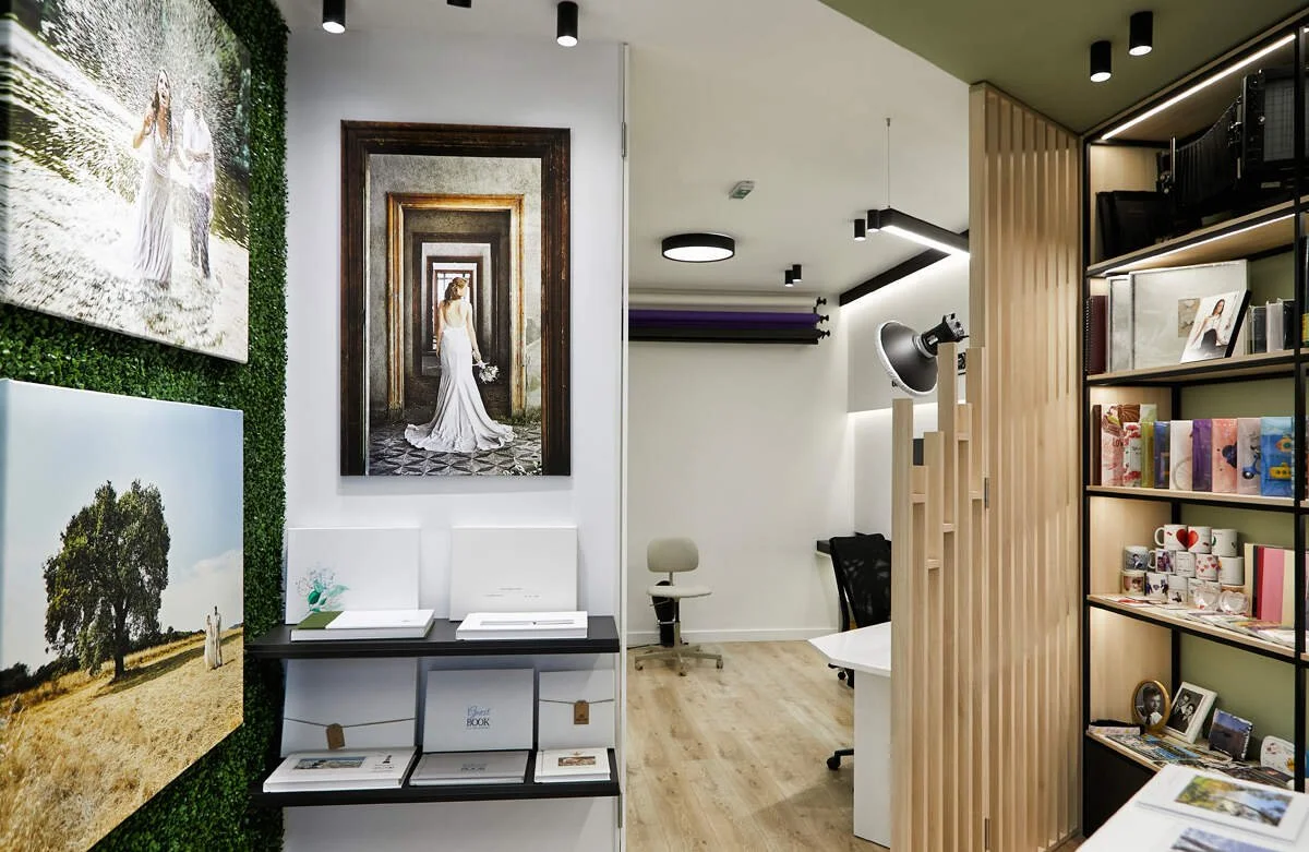 Interior of a photography studio or office with pictures of a woman in a wedding dress on the wall, shelves with books and photo prints, a large studio light, and office chairs.