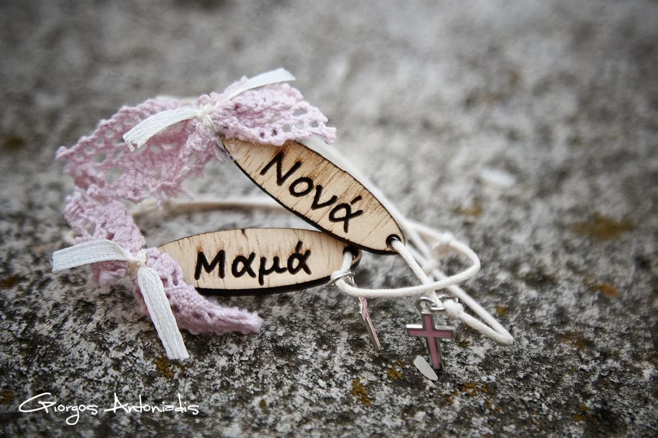 A bracelet with wooden charms that have inscriptions in Greek, one says 'Mom' and the other 'Now and'. It includes a pink cross charm, pink lace, and white ribbons, lying on a rough surface.