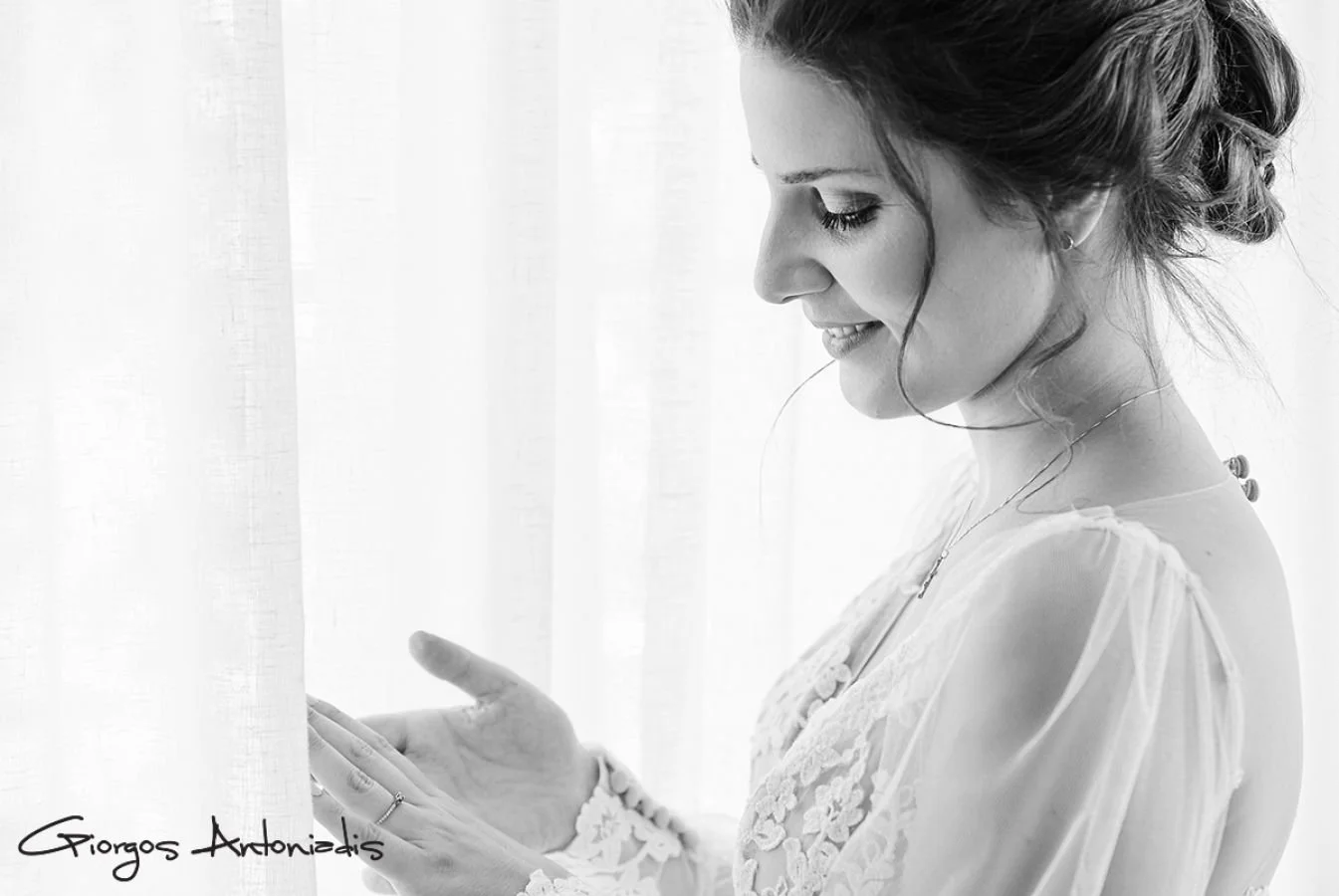 Black and white photo of a woman smiling gently, looking down at her phone, standing near a light-curtained window. She is wearing a delicate lace dress, with her hair styled in an elegant updo, and has jewelry on her wrist and neck.