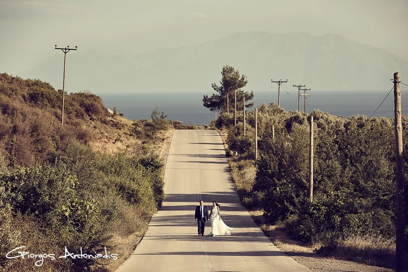 A bride and groom walking hand in hand down a deserted country road surrounded by greenery, with power lines and a mountain in the background.