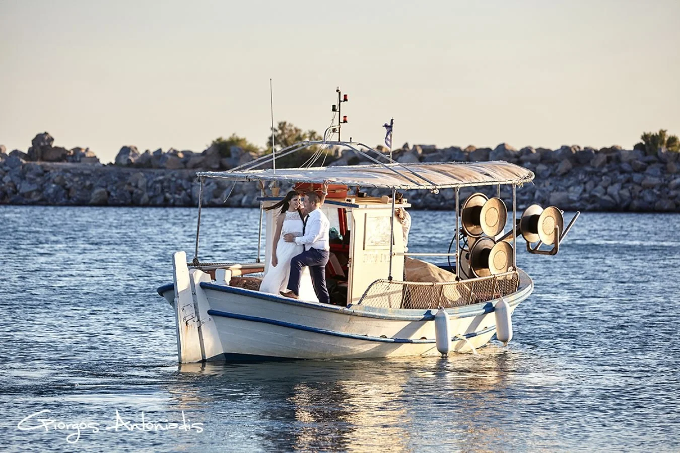 A couple in wedding attire on a small boat on a calm body of water during sunset, with a rocky shoreline in the background.