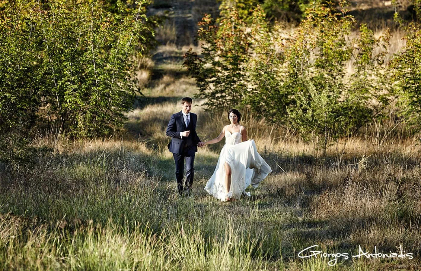 A bride and groom holding hands walking through a grassy field surrounded by trees during a sunny day.