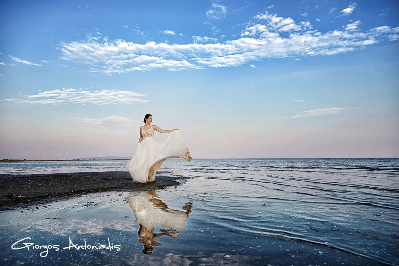A woman wearing a wedding dress on a beach, holding up the hem of her dress, with water and clouds in the sky reflected in the wet sand.