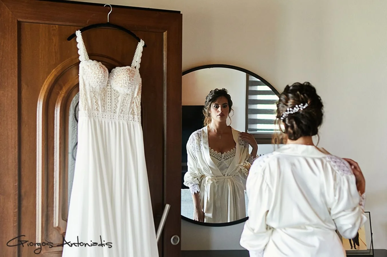 A woman in a satin robe with lace details looking at herself in a round mirror. A wedding dress hanging on a wooden door in front of her.