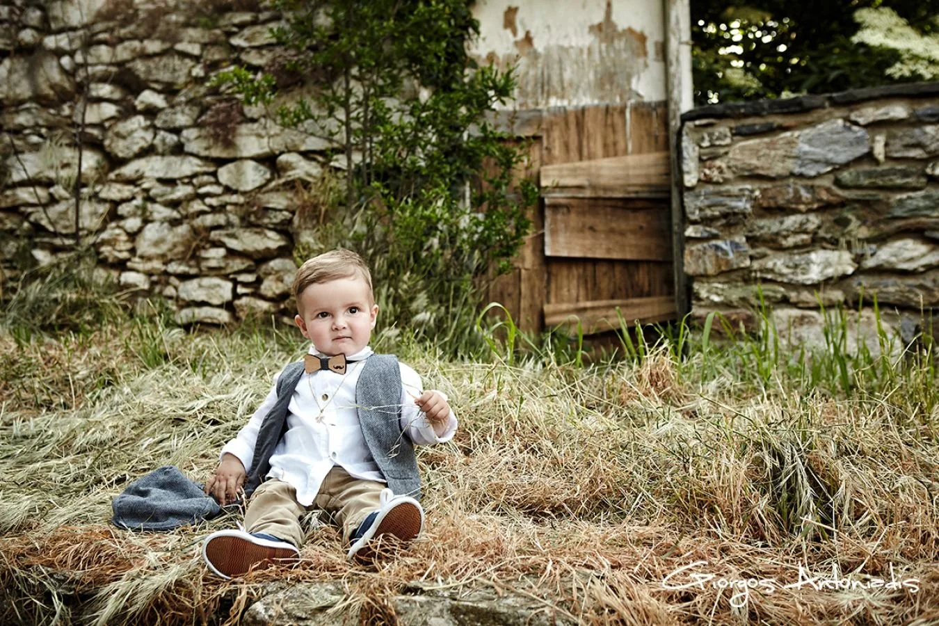 Young boy sitting outdoors on dry grass, wearing a white shirt, gray vest, beige pants, and a bow tie, with a stone wall and wooden gate in the background.