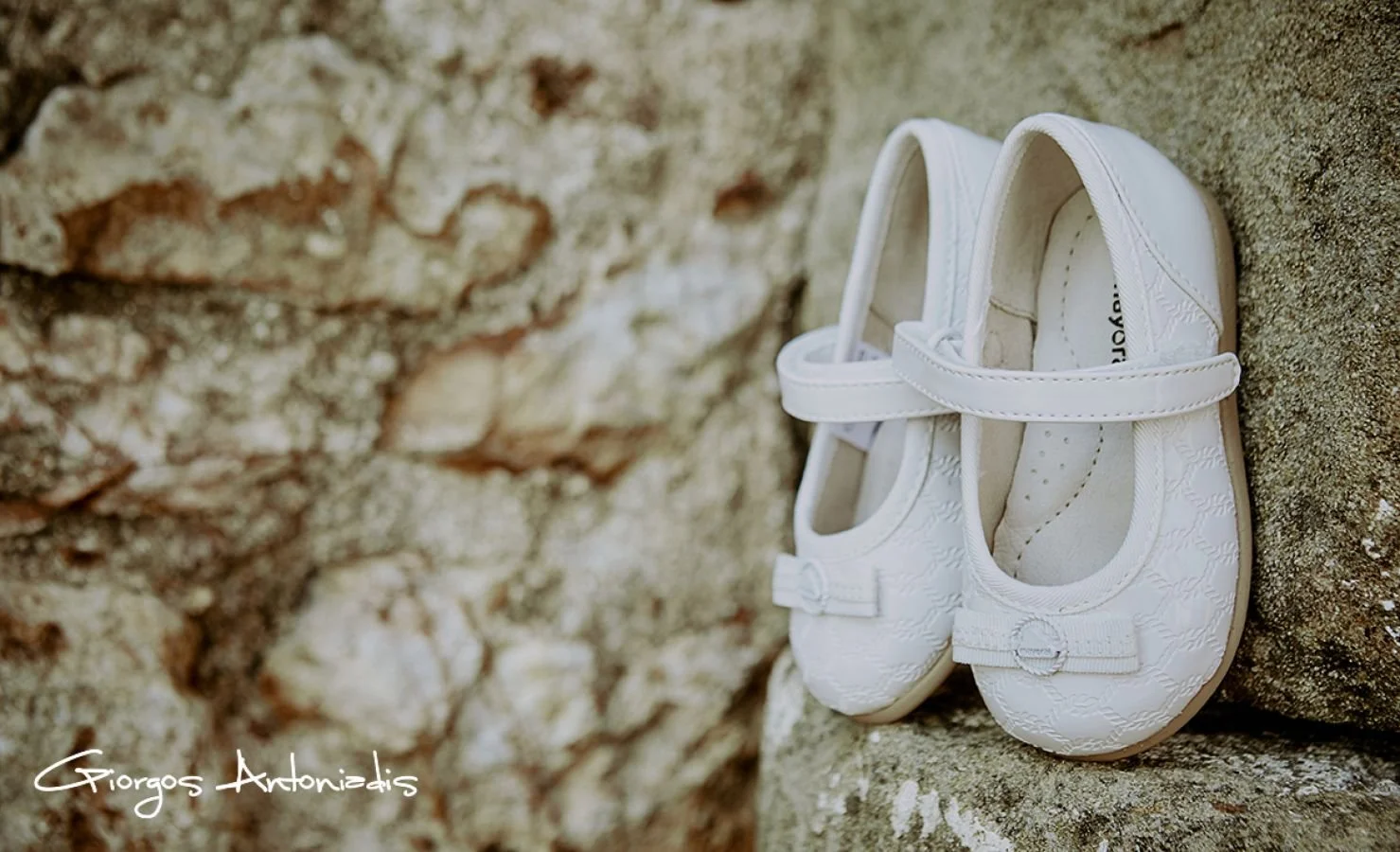 Pair of white baby shoes with a bow detail on a rough stone surface.