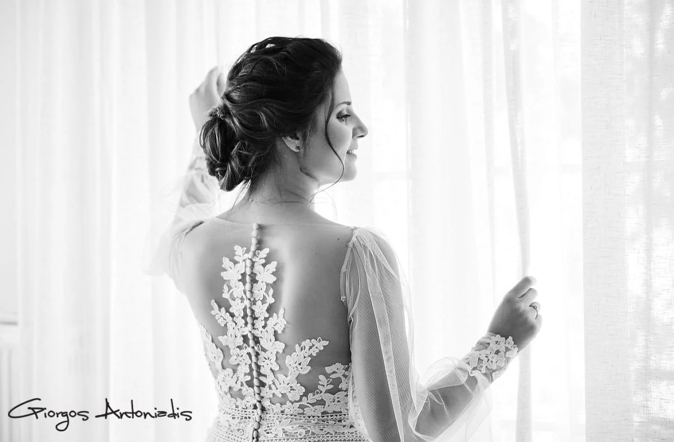 A woman with dark hair styled in an updo, wearing a lace dress with floral embroidery on the back and sheer long sleeves, standing near a window with curtains and smiling softly.