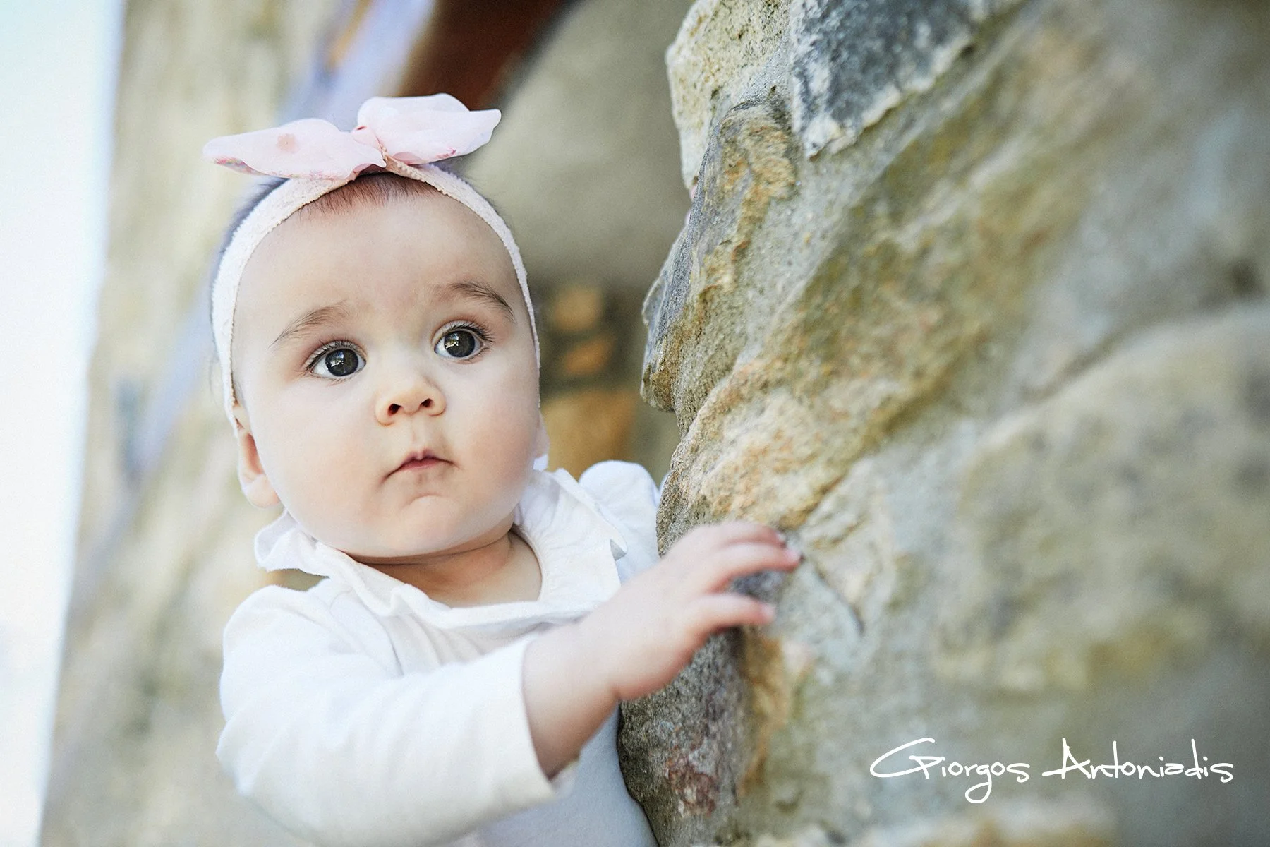 A young girl with big, expressive eyes and a pink headband with a bow, touching a rocky stone wall.