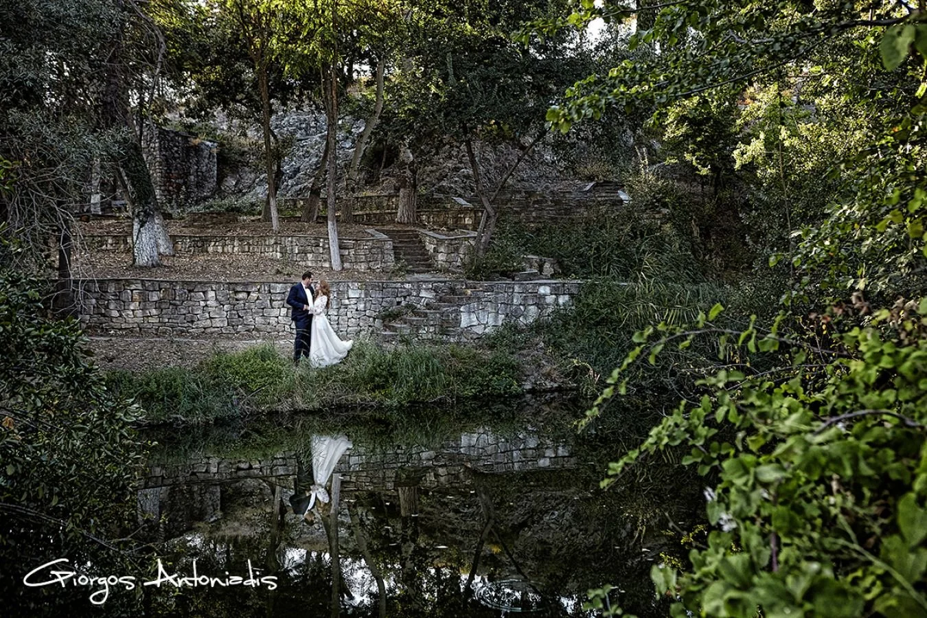 A couple in wedding attire standing by a small pond with trees and stone steps behind them, reflected in the water, in a lush outdoor setting.