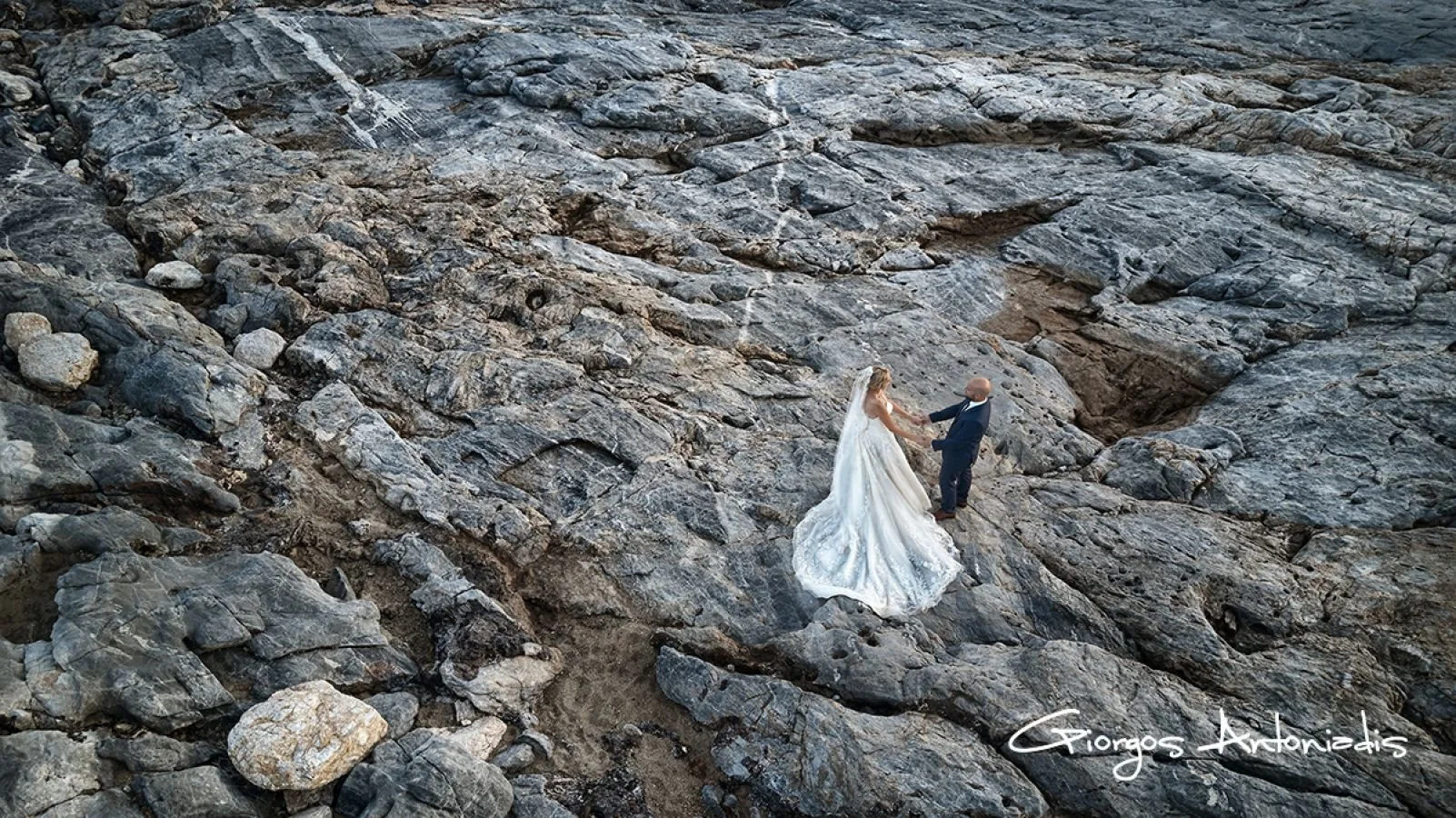 A bride and groom holding hands on rocky terrain, with the bride in a white wedding dress and veil, and the groom in a dark suit, during a wedding photoshoot