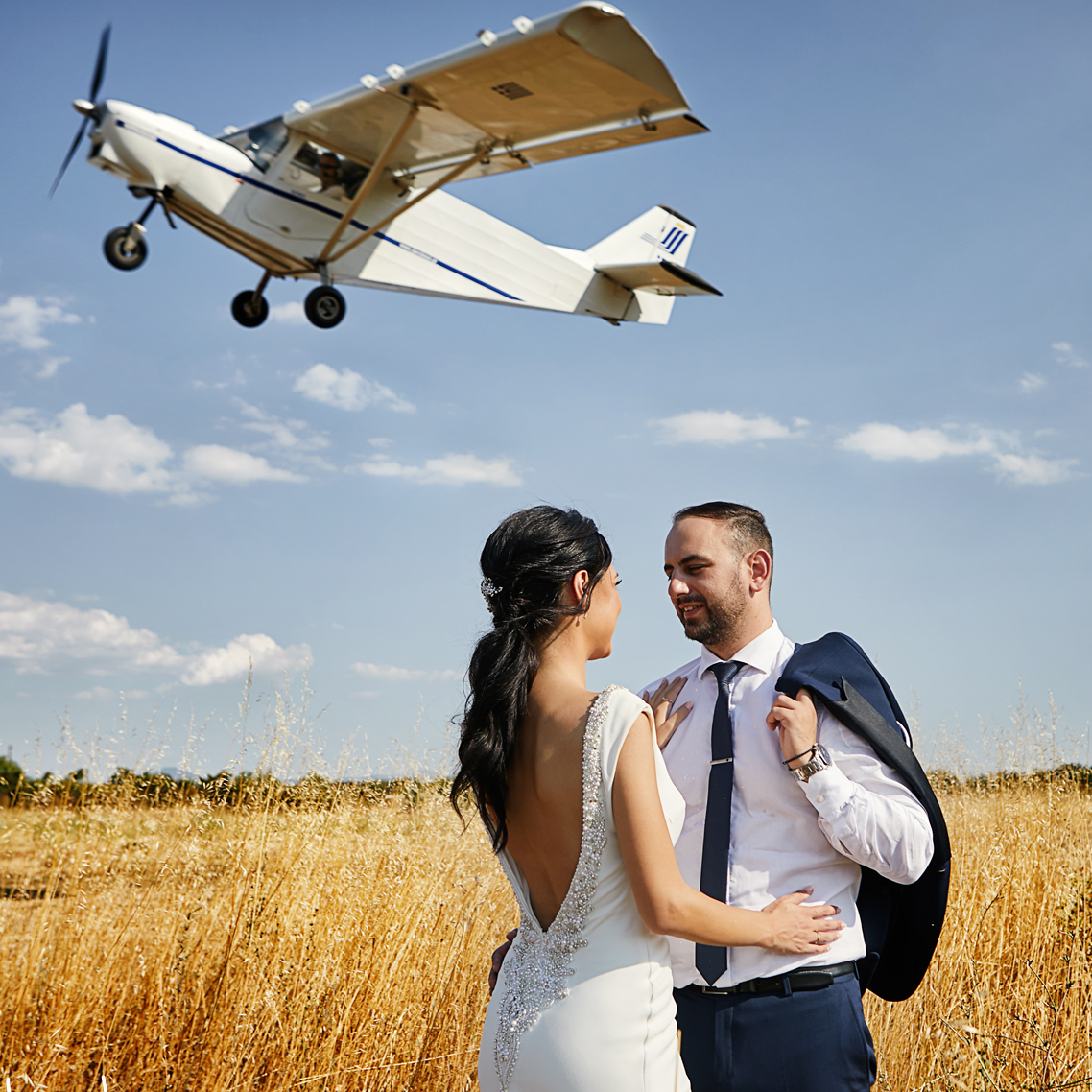 A couple in wedding attire standing in a field of tall golden grass, smiling at each other, with a small airplane flying overhead against a blue sky with some clouds.