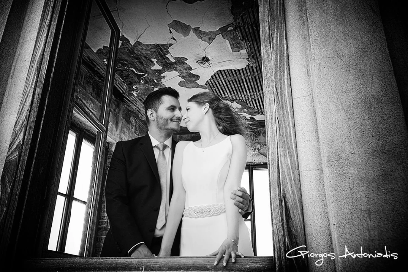 A black and white photo of a bride and groom looking at each other affectionately through a window frame. The groom has a beard and is wearing a suit with a tie, while the bride has long hair and is dressed in a sleeveless wedding gown.