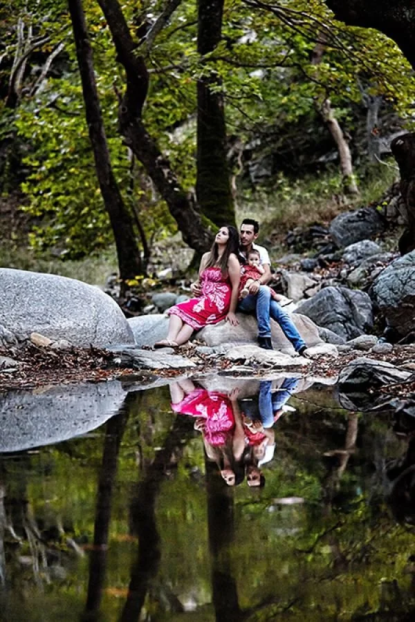 A family of three sitting on a large rock by a creek in a forested area, with their reflection visible in the water.