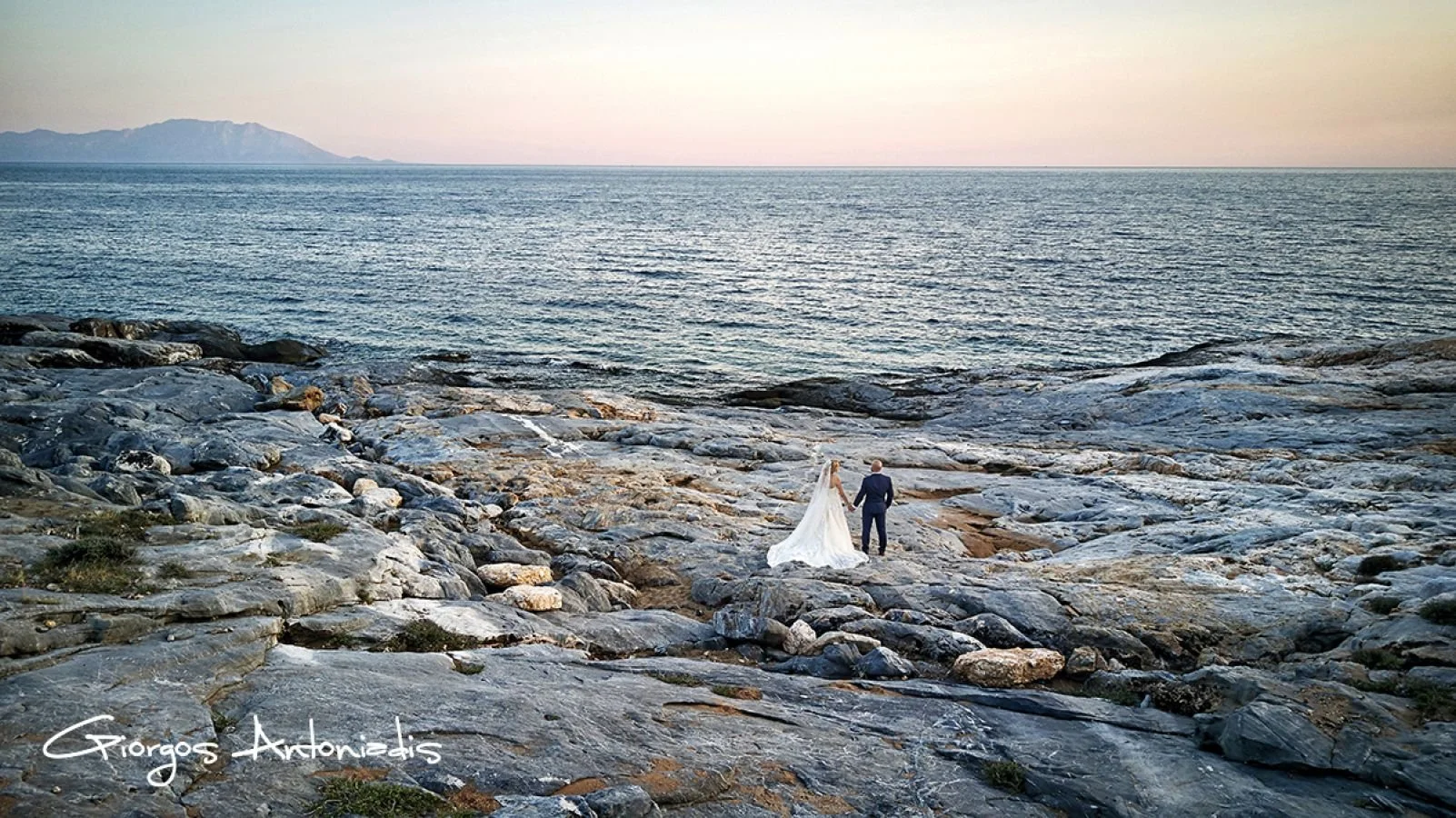 A bride and groom holding hands on a rocky shoreline with the ocean and mountains in the background.