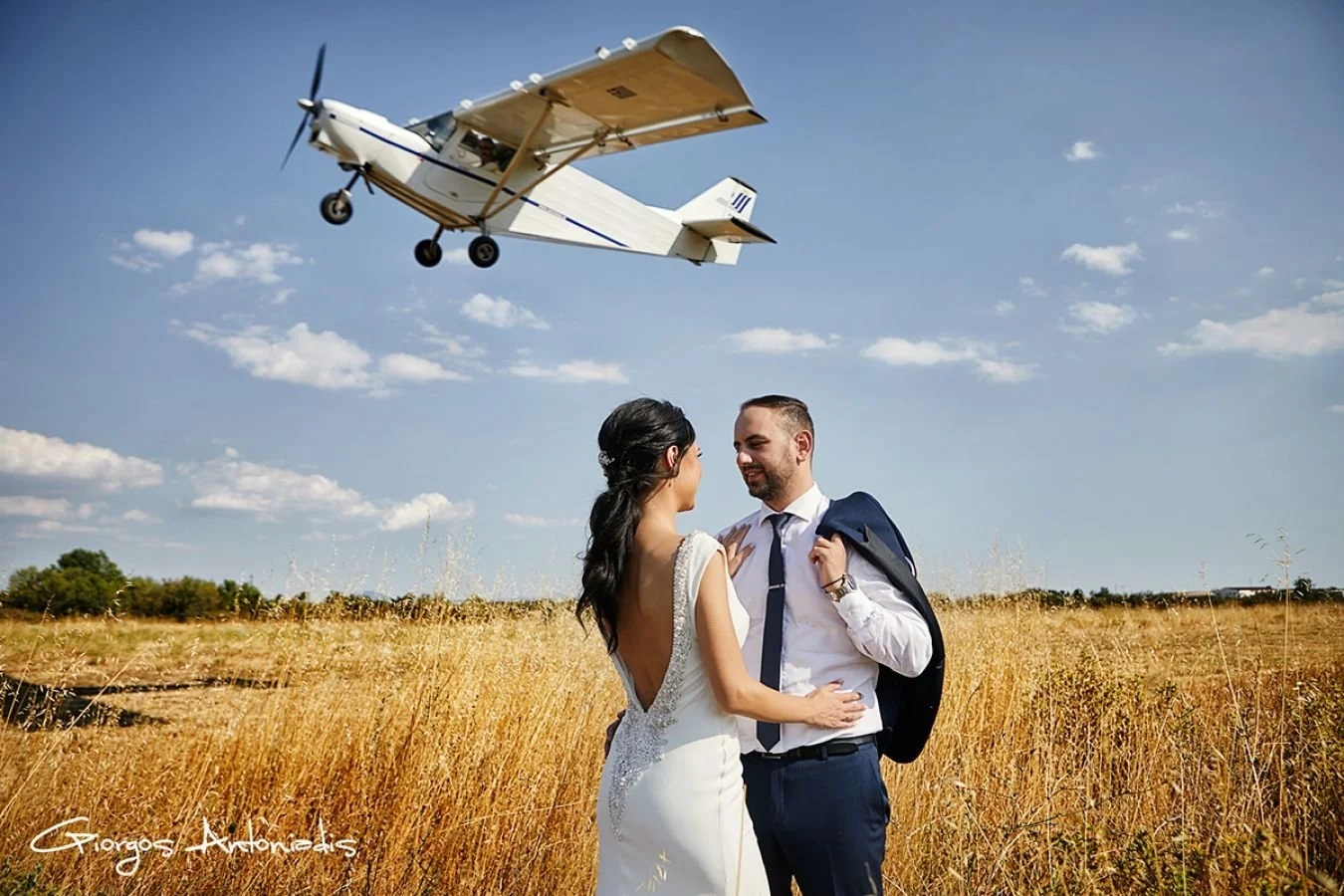 A couple, dressed in wedding attire, standing in a field and smiling at each other as a small airplane flies overhead on a sunny day with a blue sky.
