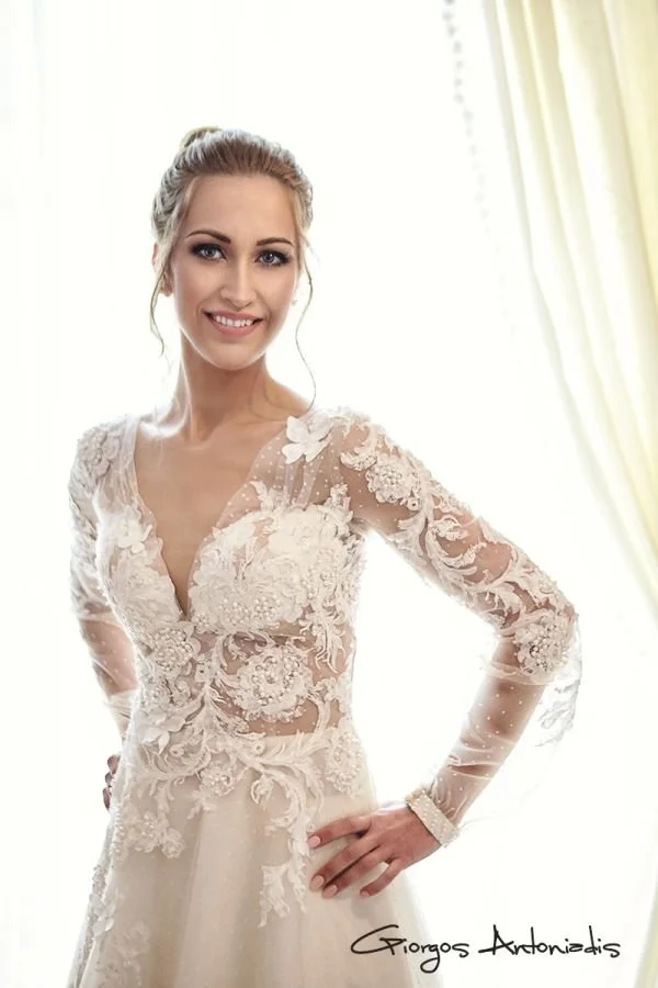 A smiling woman in a wedding dress with lace details, standing indoors near light-colored curtains.
