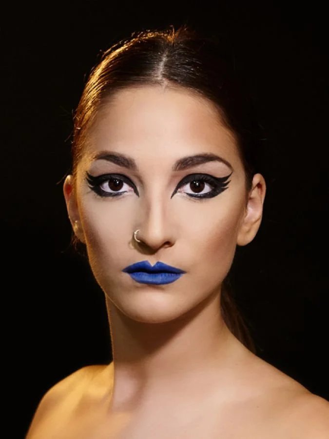 Close-up portrait of a woman with bold makeup and blue lipstick, black winged eyeliner, and a nose ring, against a dark background.