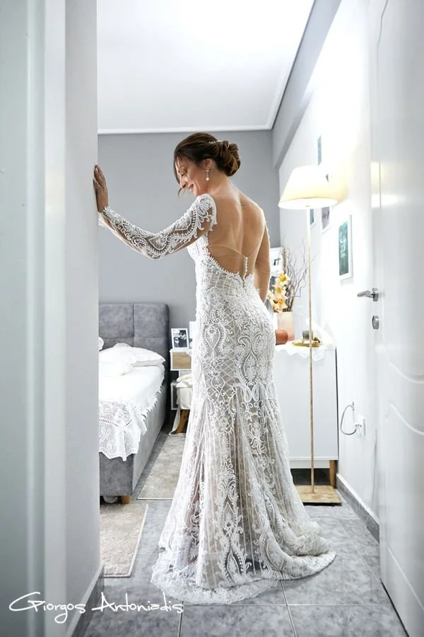 A bride in a white lace wedding gown standing in a bedroom, smiling and looking down, with a bed, side table, and art on the walls in the background.