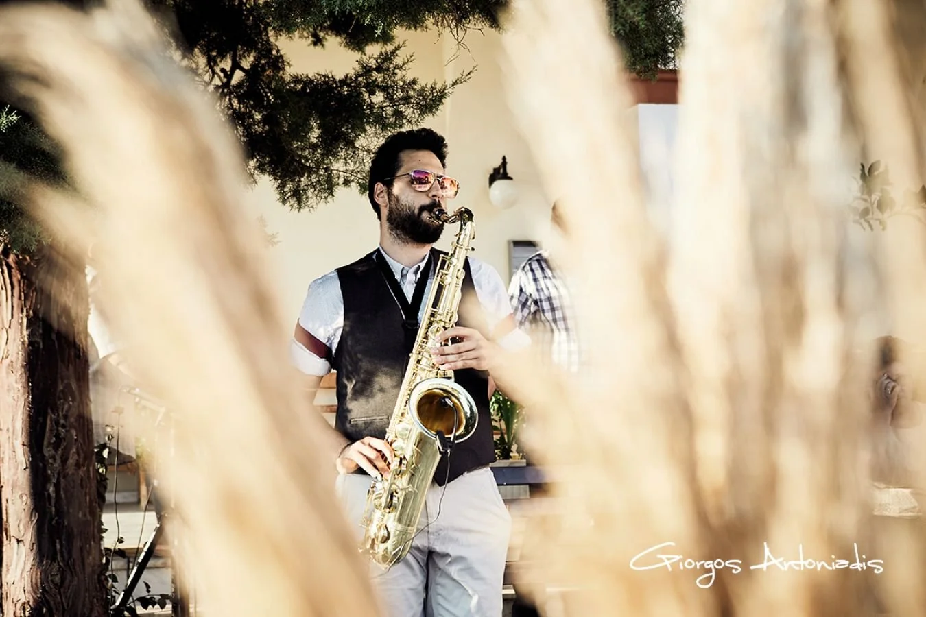 Man with sunglasses playing saxophone outdoors, framed by tree branches, signature 'Giorgos Antonidis' in bottom right corner.