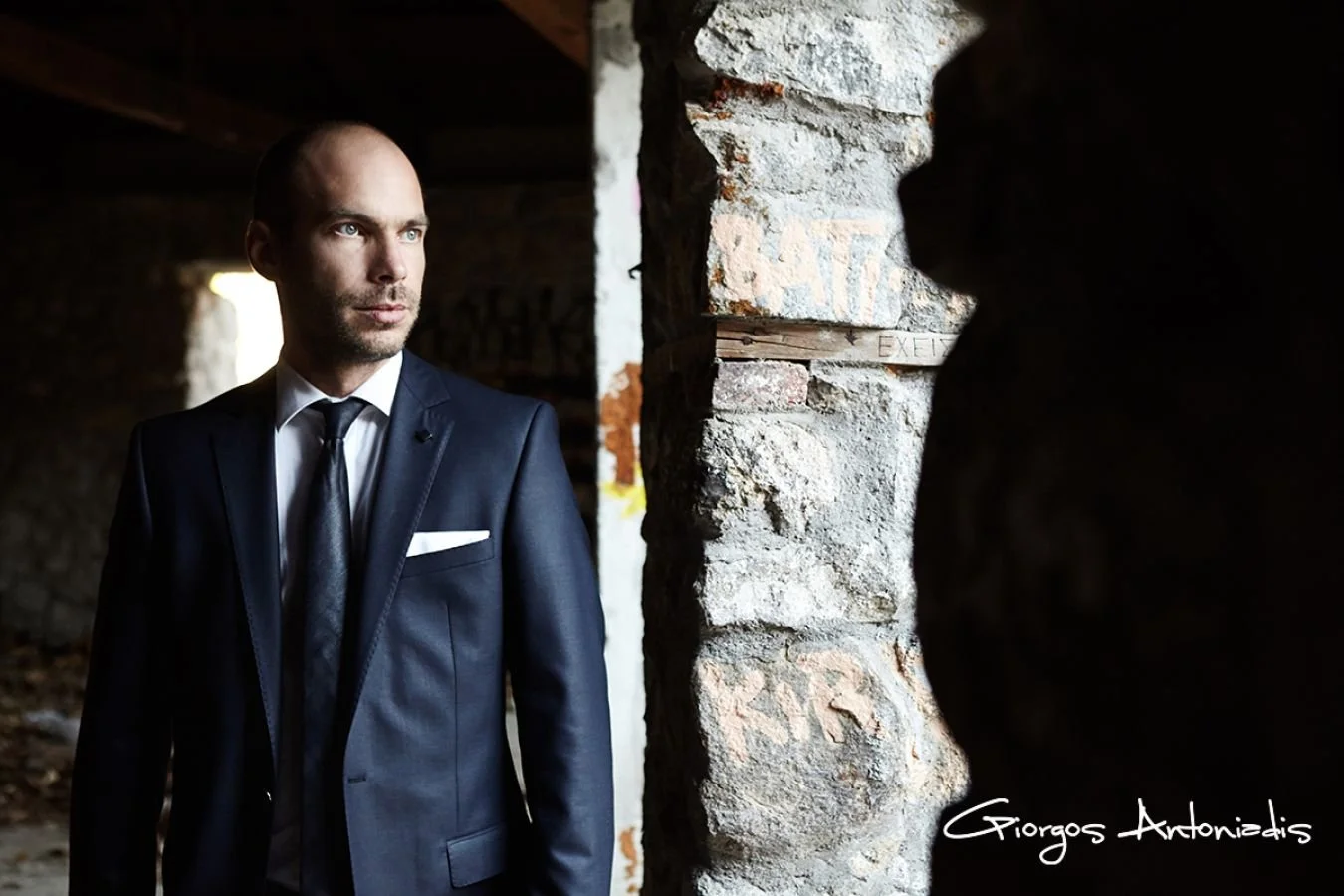 A man in a business suit standing near a stone wall in an underground or abandoned building, looking thoughtfully into the distance.