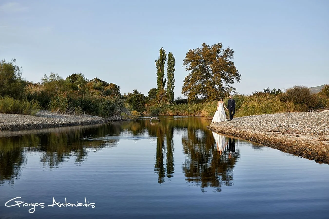 A bride and groom walking on a rocky riverbank near calm water, with trees and bushes in the background, reflecting their image in the water, during sunset or early evening.