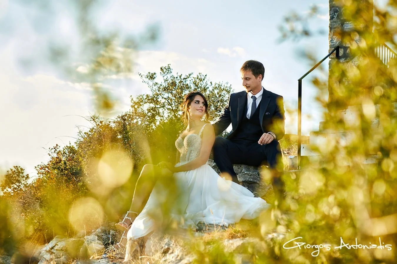 A couple dressed in wedding attire sitting on rocks outdoors surrounded by foliage and trees, during daylight.