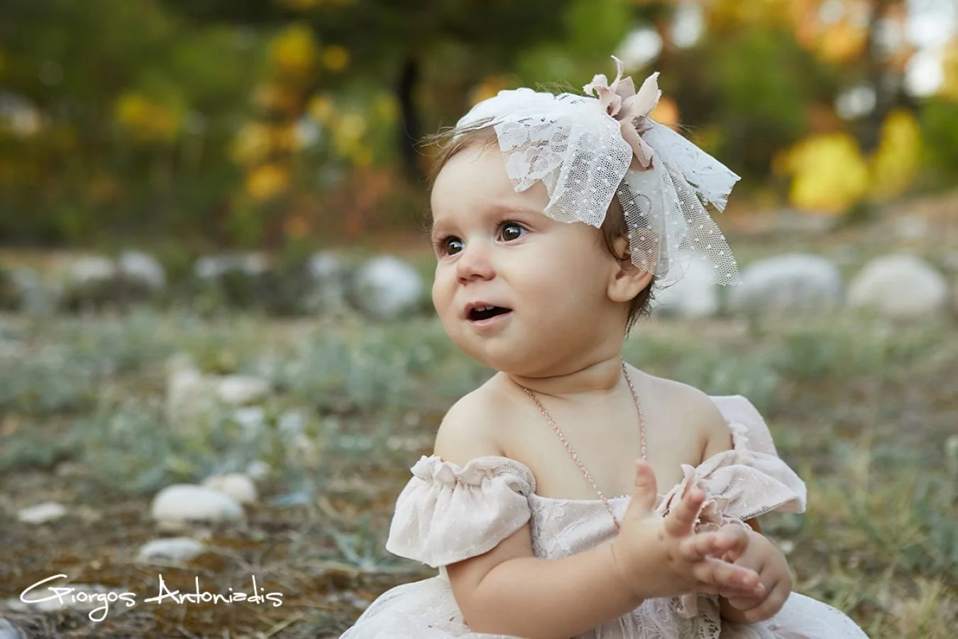 A young child in an off-shoulder dress with puffed sleeves, wearing a lace headband with a bow, sitting outdoors on the grass with autumn trees in the background.