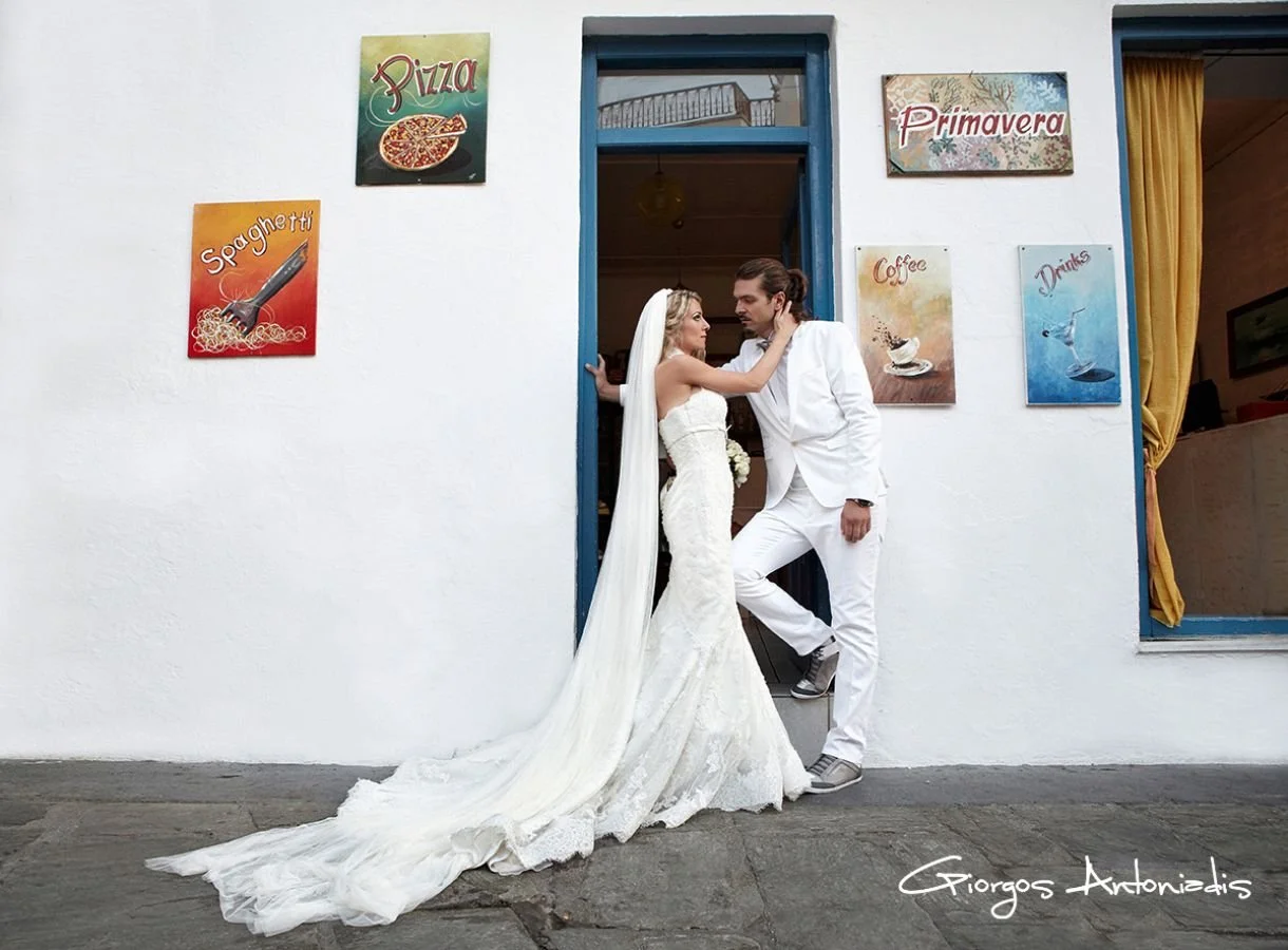 A bride and groom in wedding attire pose in front of a white wall with various colorful signs advertising pizza, spaghetti, primavera, coffee, and drinks. The bride wears a white wedding gown with a long train, and the groom wears a white suit. They 