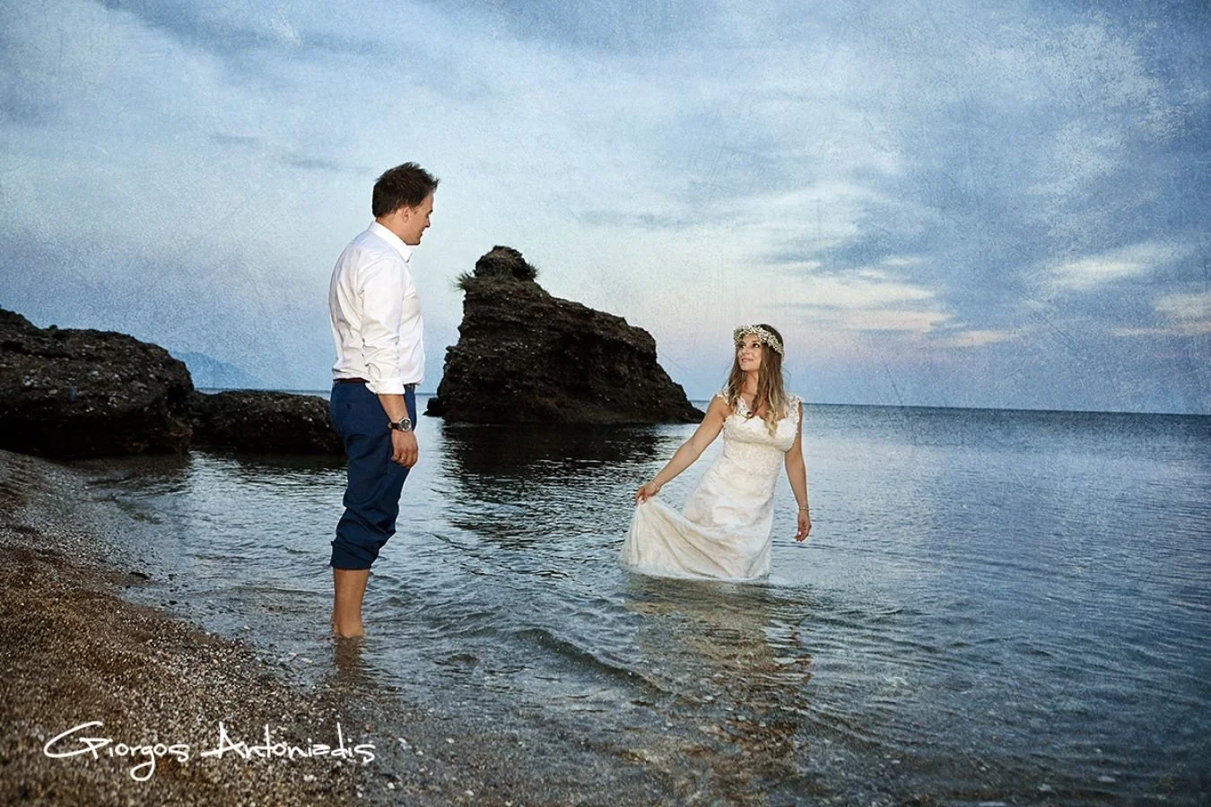 A man and a woman stand in shallow ocean water on a rocky beach, with the woman in a white lace dress and floral crown, and the man in a white shirt and blue pants. The woman is holding her dress while looking at the man, with large rocks and a cloud