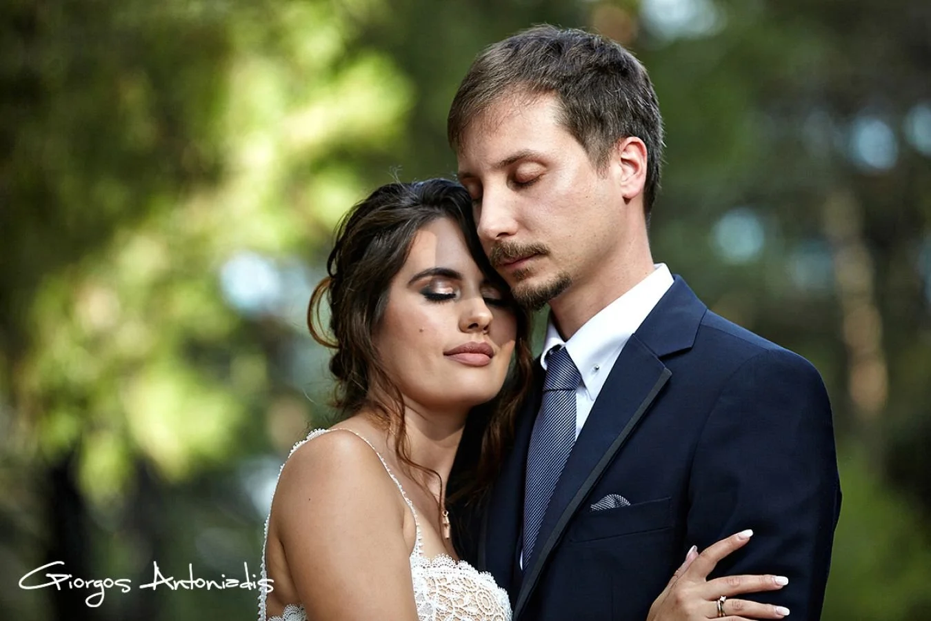 A couple embracing each other outdoors, with their eyes closed, dressed in formal attire, surrounded by a blurred green background.
