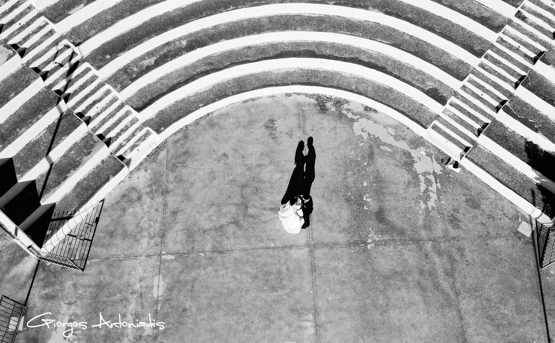 Black and white aerial photo of a person standing on a concrete ground near a set of curved stairs, with their shadow cast on the ground.