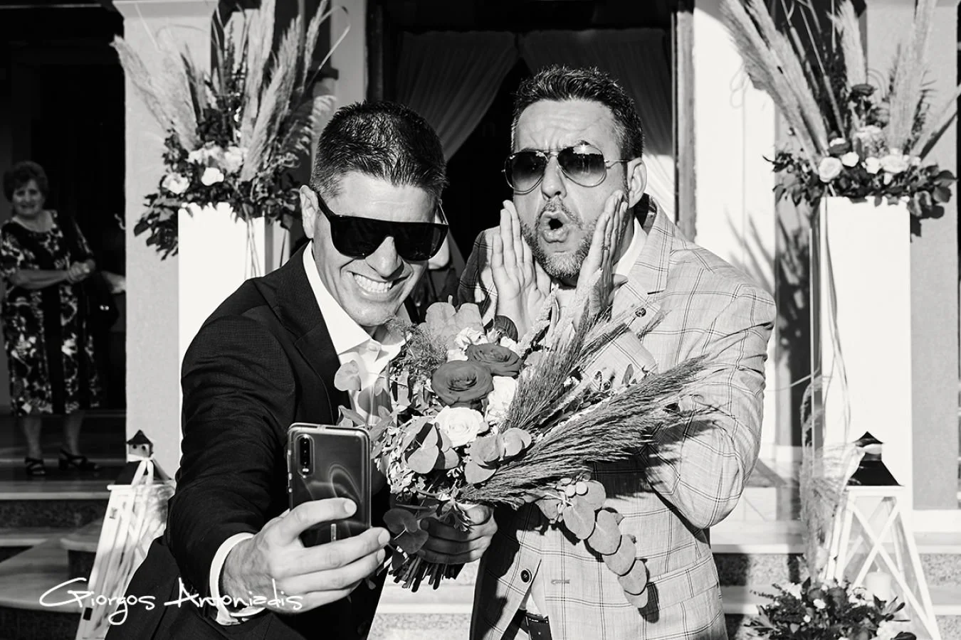 Two men wearing sunglasses and smiling, one taking a selfie with a smartphone, holding a bouquet of flowers, at a celebration or wedding event.