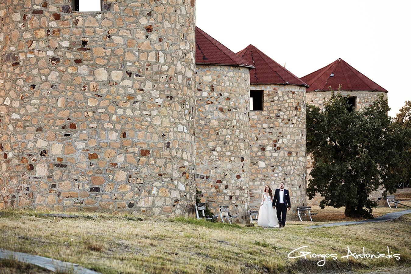 A bride and groom holding hands walking near a stone castle with red roofs, trees, and benches in the background.