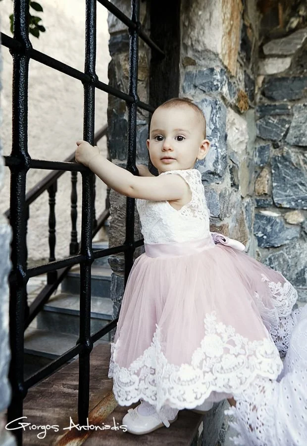 A young girl dressed in a white lace top and a pink tulle skirt with white floral embroidery, holding onto black metal bars on a staircase near a stone wall.