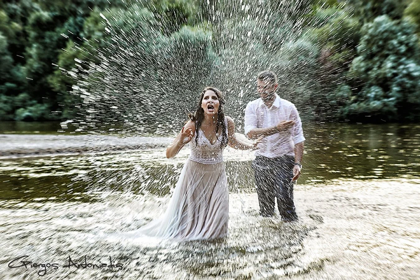 A woman in a white dress and a man in a white shirt standing in a river, being splashed with water, with trees in the background.