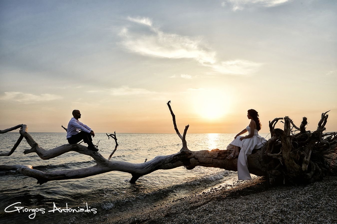 A man and woman sit on opposite ends of a large driftwood log by the ocean at sunset. The man on the left is dressed in black and white, and the woman on the right is dressed in a white dress. The sky is partly cloudy with the sun setting on the hori