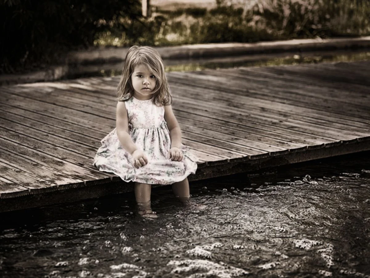 A young girl sitting on the edge of a wooden dock with her feet in the water, looking at the camera with a serious expression.
