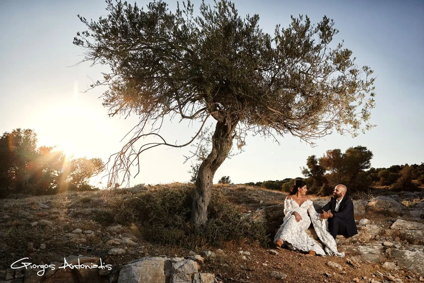 A man and woman sitting on rocky ground under a tree at sunset, the woman in a white lace dress and the man in a dark suit, holding hands and looking at each other.