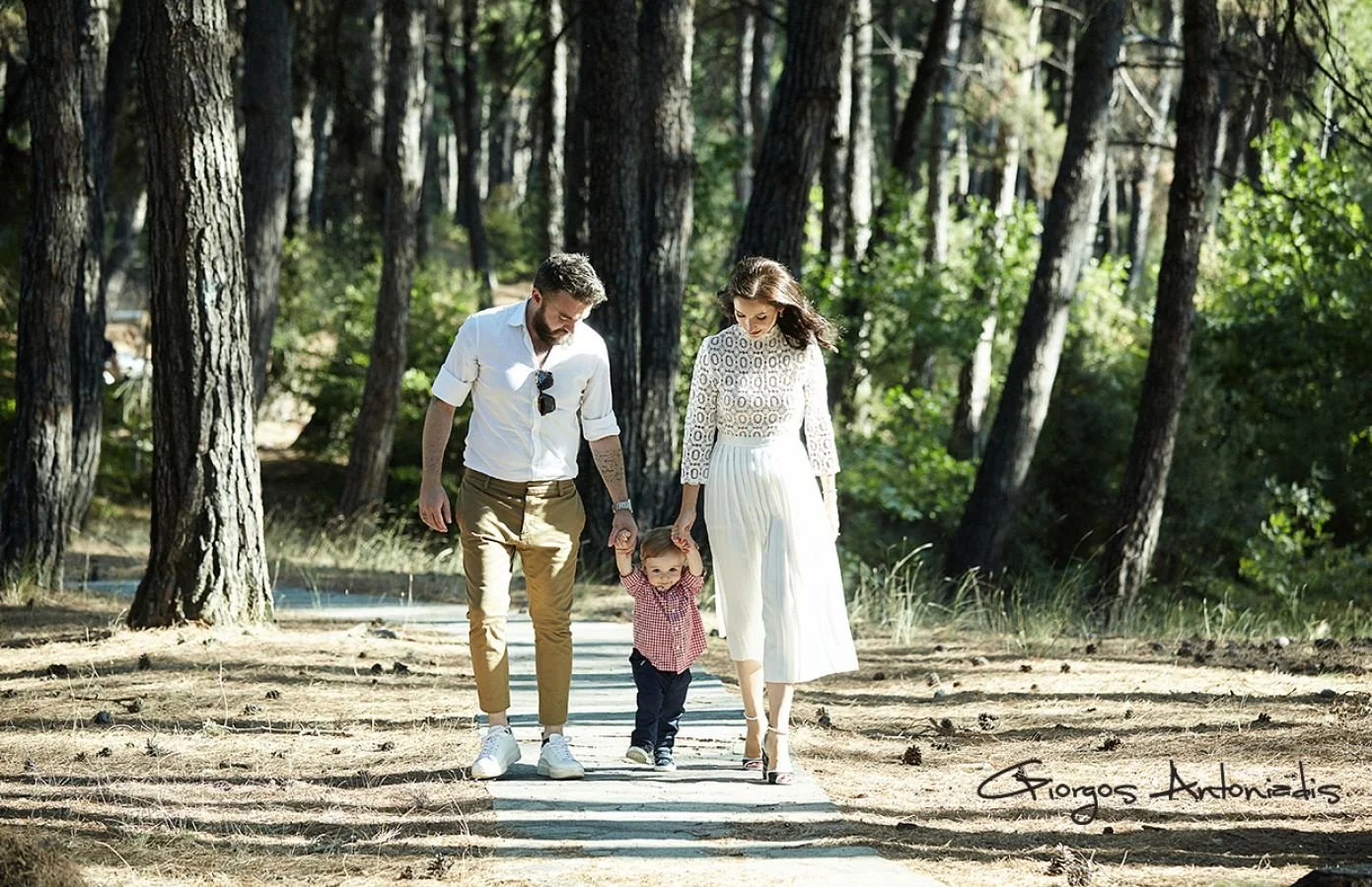 A family of three walking through a forest, holding hands. The man and woman are adults, and a young child is between them. The forest is filled with tall trees, and the sun is shining.