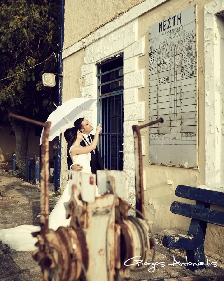 A bride and groom embrace under a white umbrella on a sidewalk in front of a building with a large metal sign and a rusty piece of equipment in the foreground.