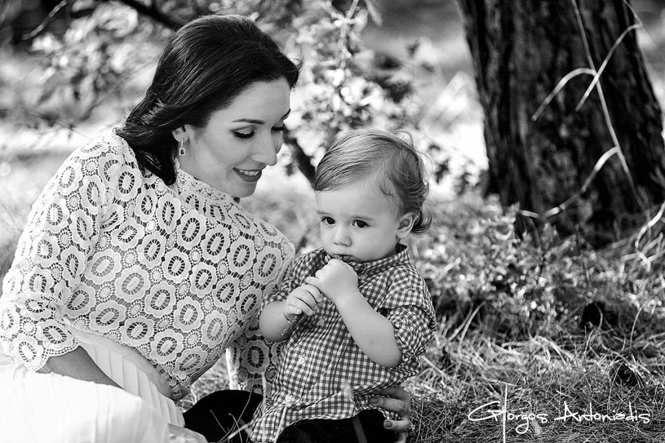 A woman and a young boy sitting outdoors, with the woman smiling and the boy looking contemplative, near a tree on a grassy area.