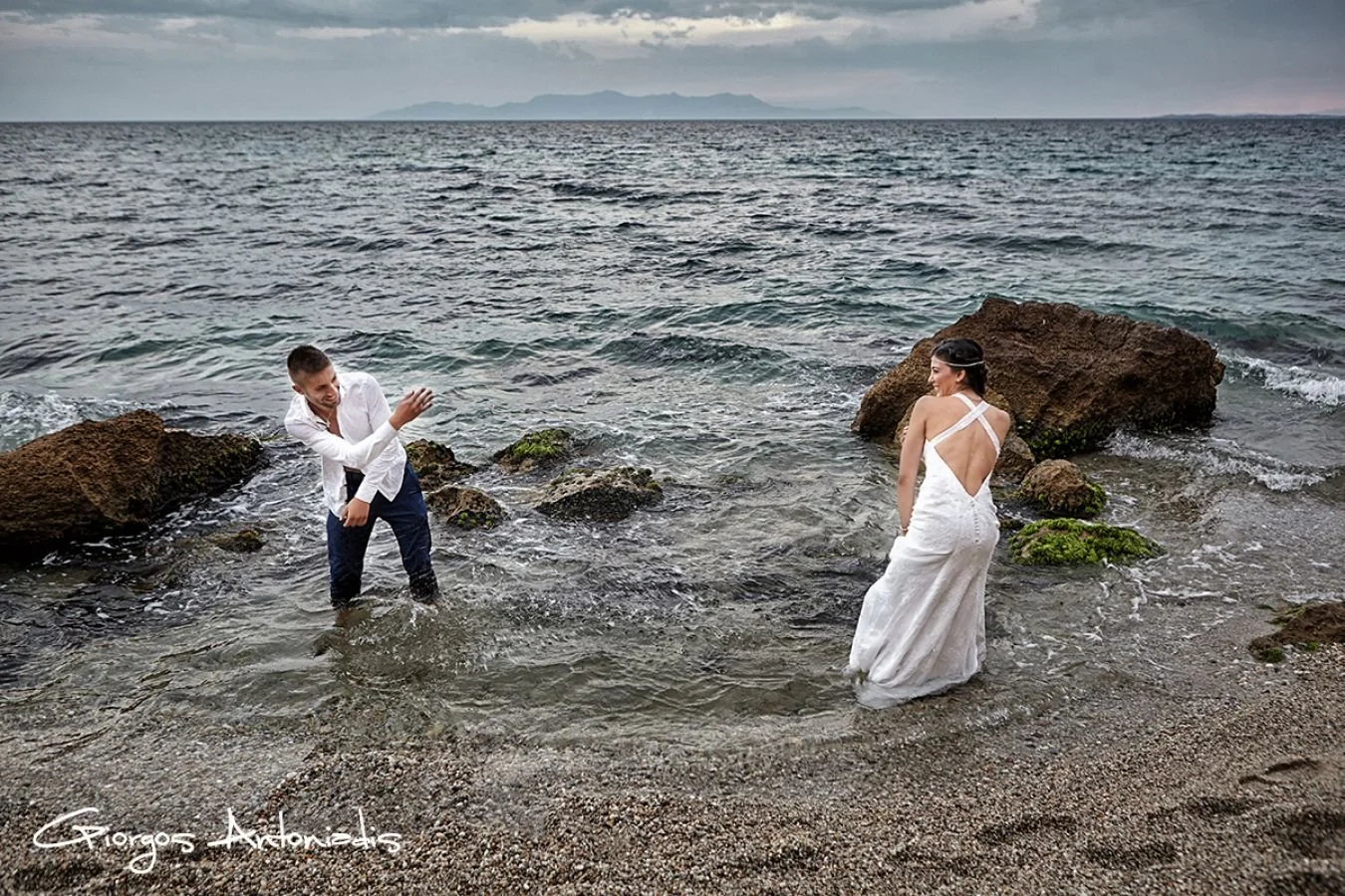 A couple dressed in wedding attire standing in the shallow waters of a beach, with rocks around them and a distant mountain range in the background.