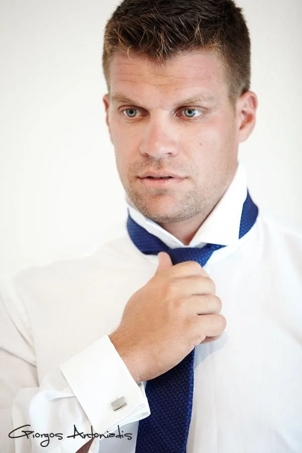 A man with short brown hair and blue eyes adjusting a blue tie while wearing a white dress shirt.