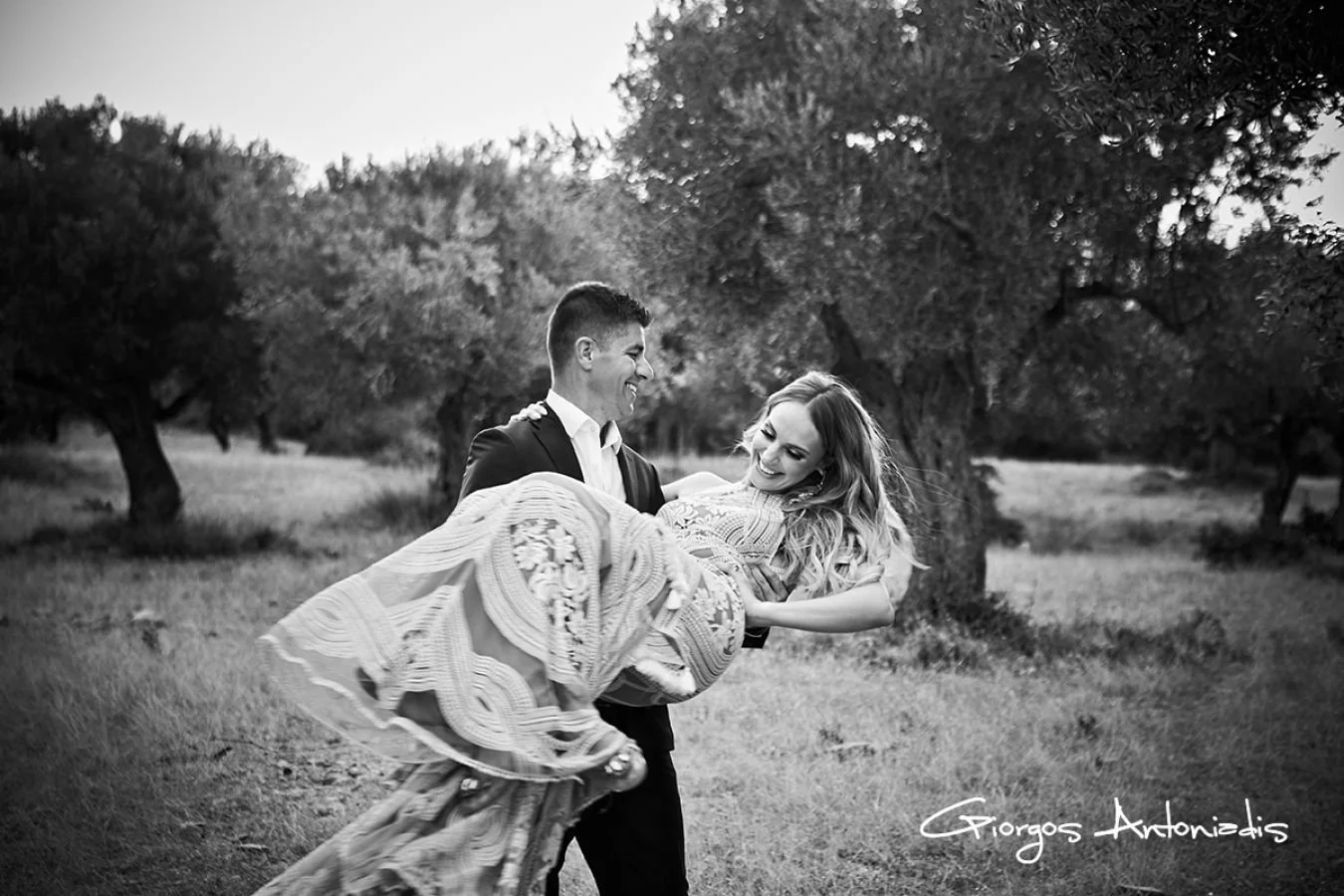 A man in a suit holding a woman in a dress in a field with trees, smiling at each other, in black and white.