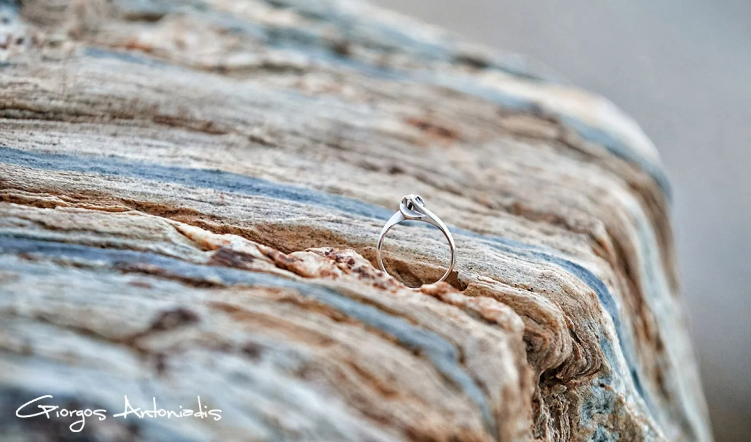 A silver ring with a small gemstone resting on a textured, layered piece of wood with shades of brown and blue.